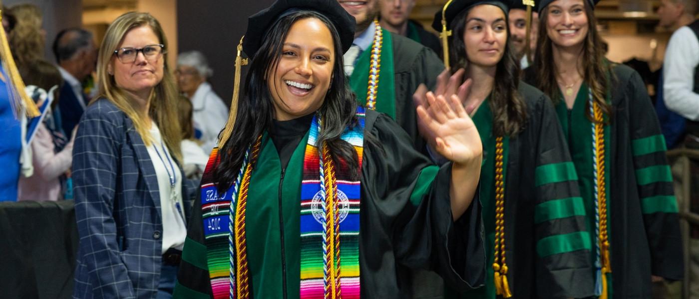 A UNE graduate wearing doctoral regalia waves walking into the ceremony